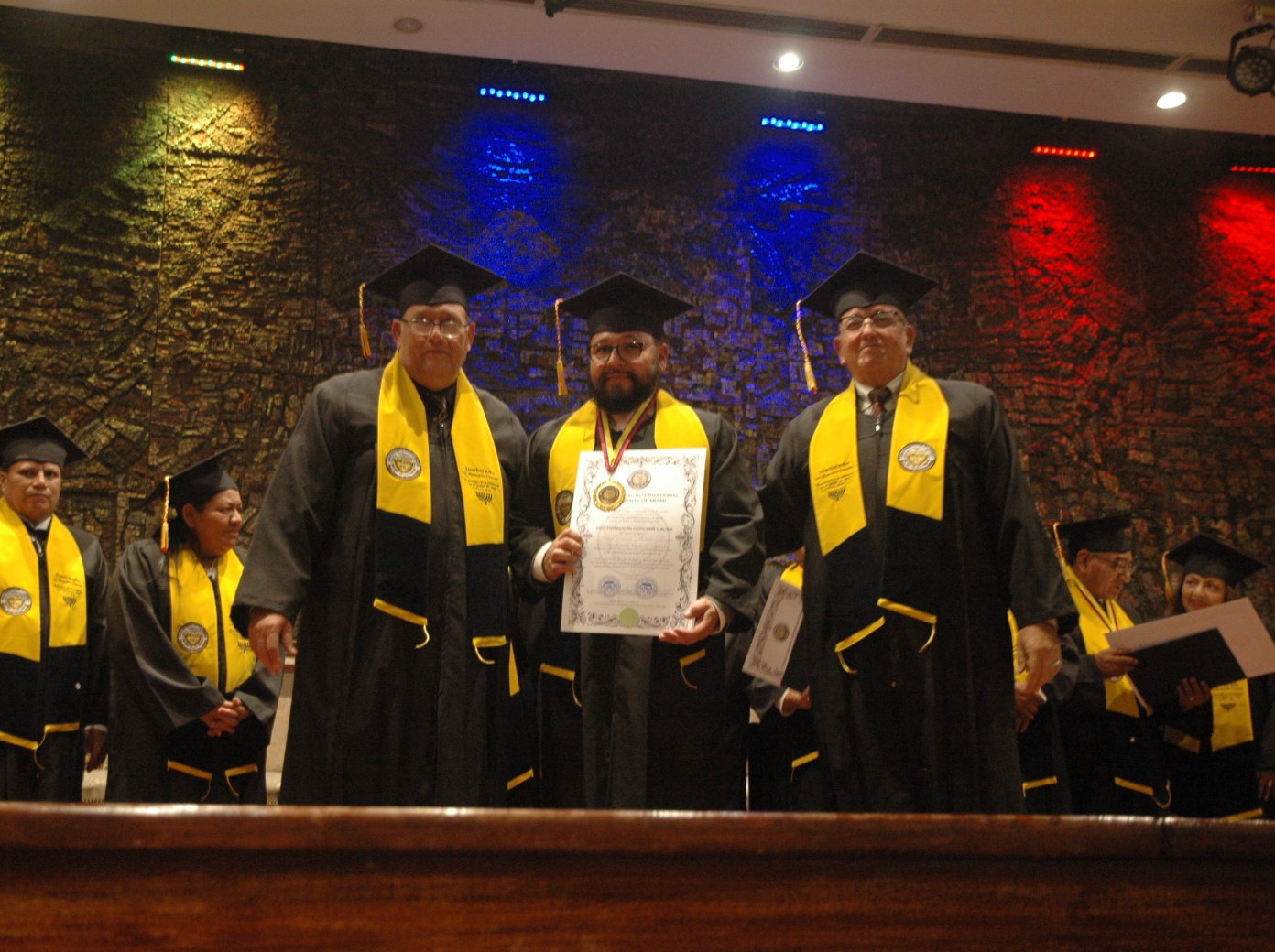 Entrega de un reconocimiento TIU Miami en el escenario a un graduado con toga y diploma